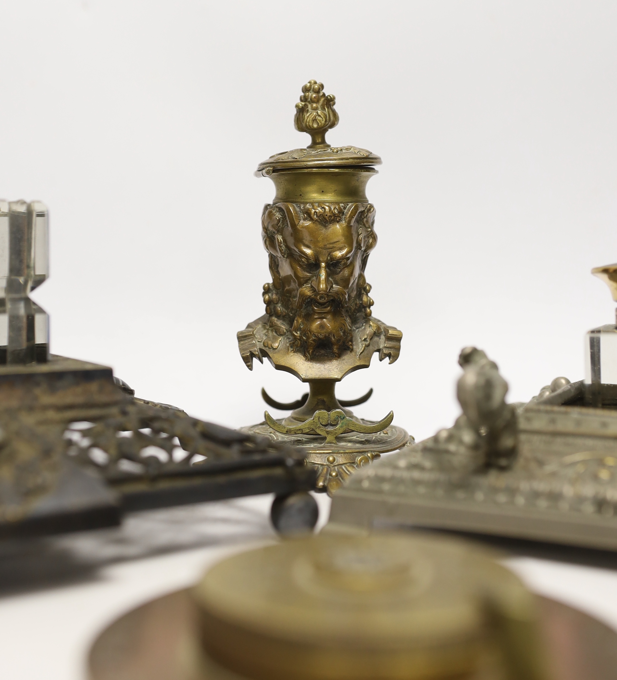Three cast metal and cut glass ink stands, including a bronze inkwell in the form of the head of Bacus, and a brass calendar display from the Banca Commerciale Italiana, London Branch, tallest 13cm